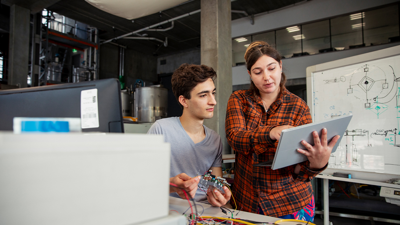 A front view of two classmates or workmates looking at a digital tablet and helping each other out with work in their on-site geothermal engineering plant in Perth, Western Australia.