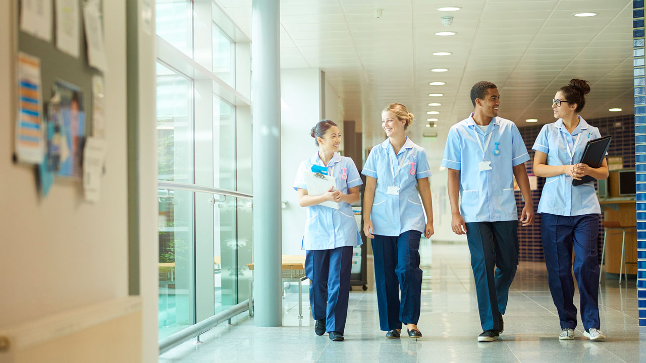 a group of four young trainee nurses including male and female nurses , walk towards camera down a hospital corridor .